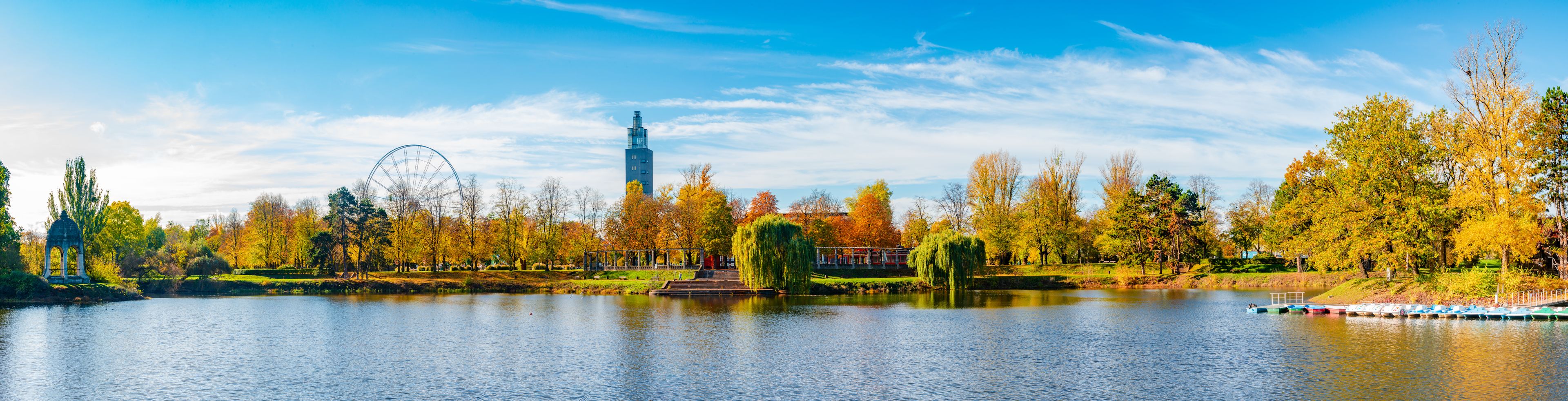 A lake with boats, autumn trees, a park with a gazebo, and a Ferris wheel.