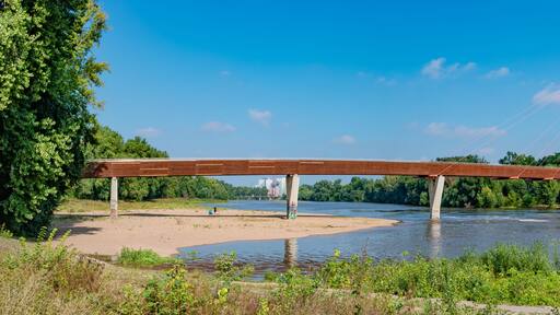 Eine rote Hängebrücke über einem Fluss, umgeben von Grünflächen und Gebäuden.