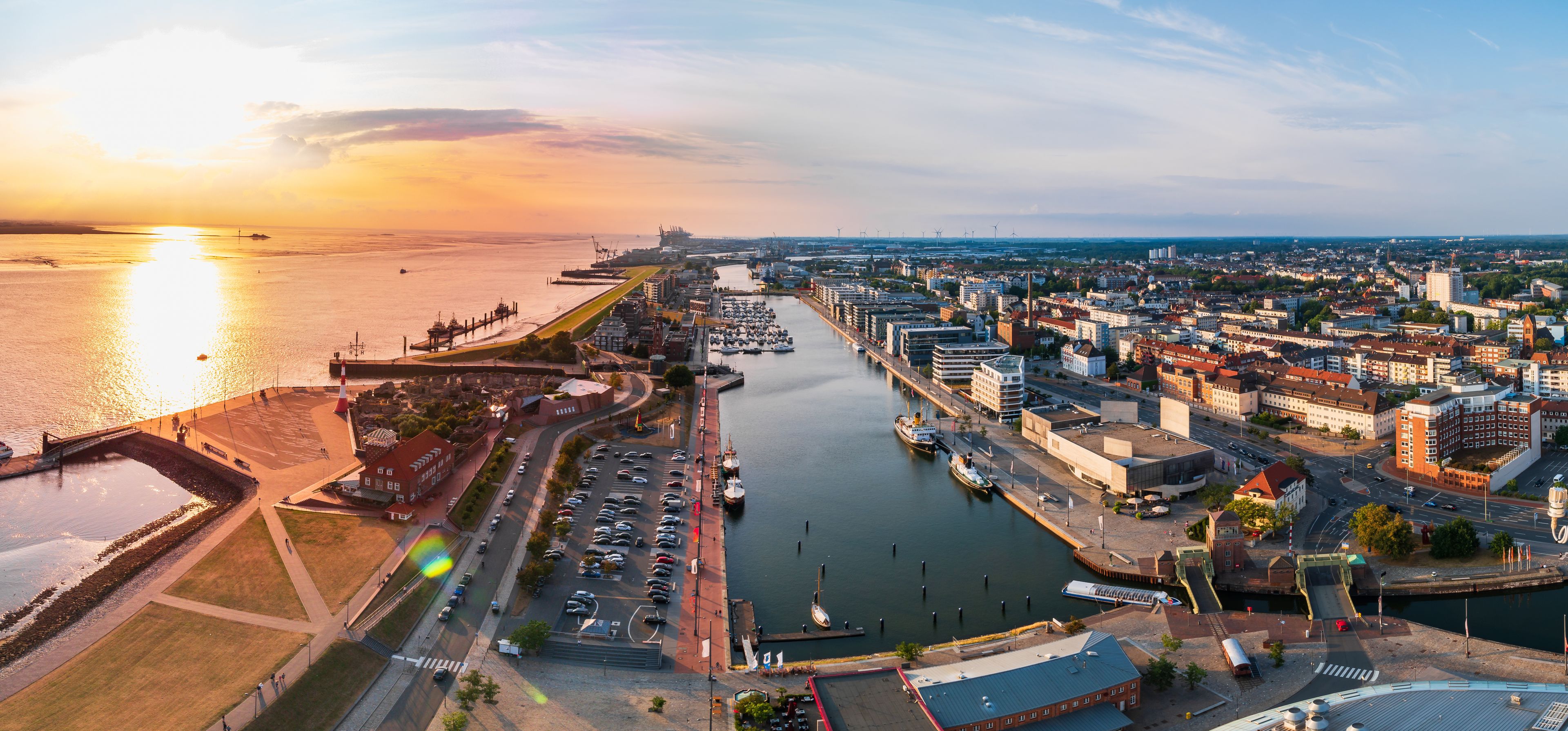 A coastal cityscape with a marina, a lighthouse, and a bridge during sunset.