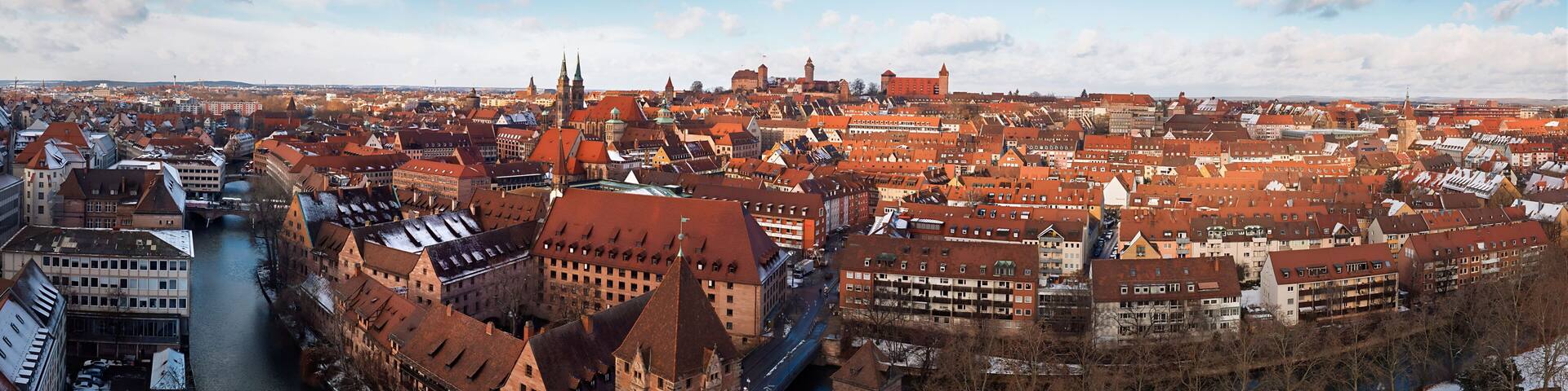 A panoramic view of a historic city with a prominent river, a castle, and a dense cluster of red-roofed buildings.