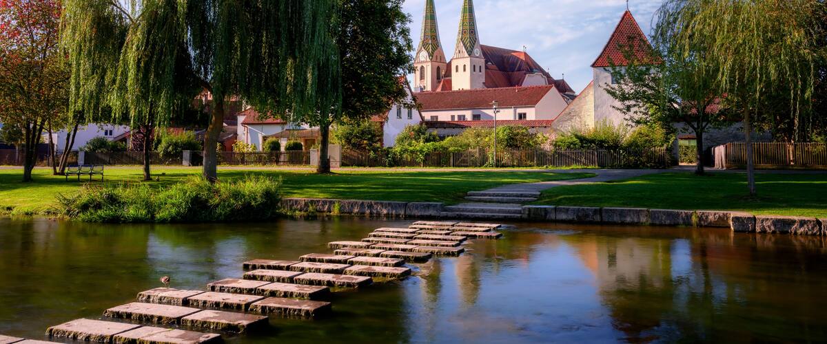Ein friedlicher Park mit einem Teich, trittsteinen, Weidenbäumen und einer Kirche mit zwei Türmen.