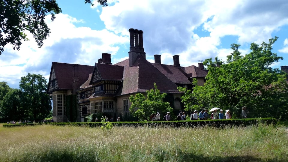 Um edifício histórico grande com telhado íngreme e várias chaminés, cercado por vegetação exuberante e um gramado bem cuidado.