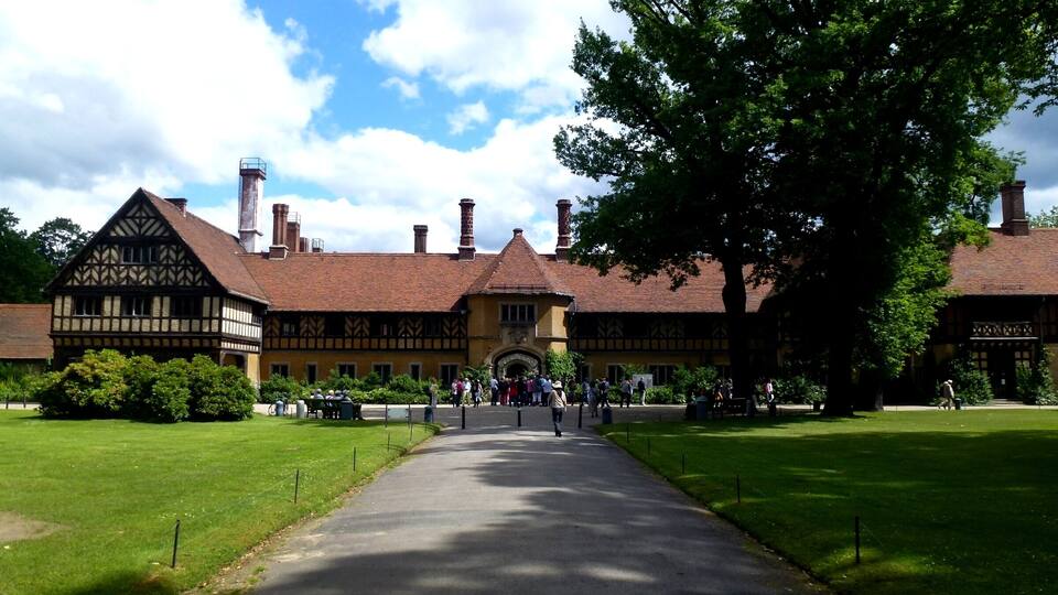 Um edifício em estilo Tudor com um arco central, rodeado por jardins bem cuidados e um caminho pavimentado que leva a ele.