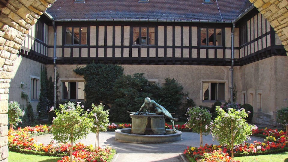 Um edifício em estilo Tudor com um pátio, uma fonte e canteiros de flores vibrantes.