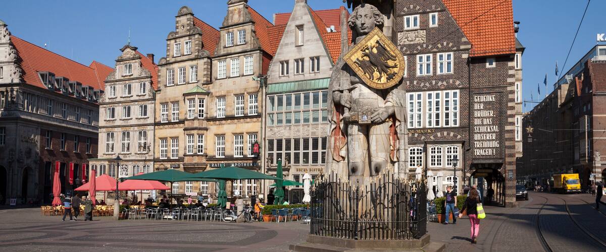 Ein historischer Platz mit traditioneller europäischer Architektur, einer markanten Statue und einer Straßenbahnhaltestelle.