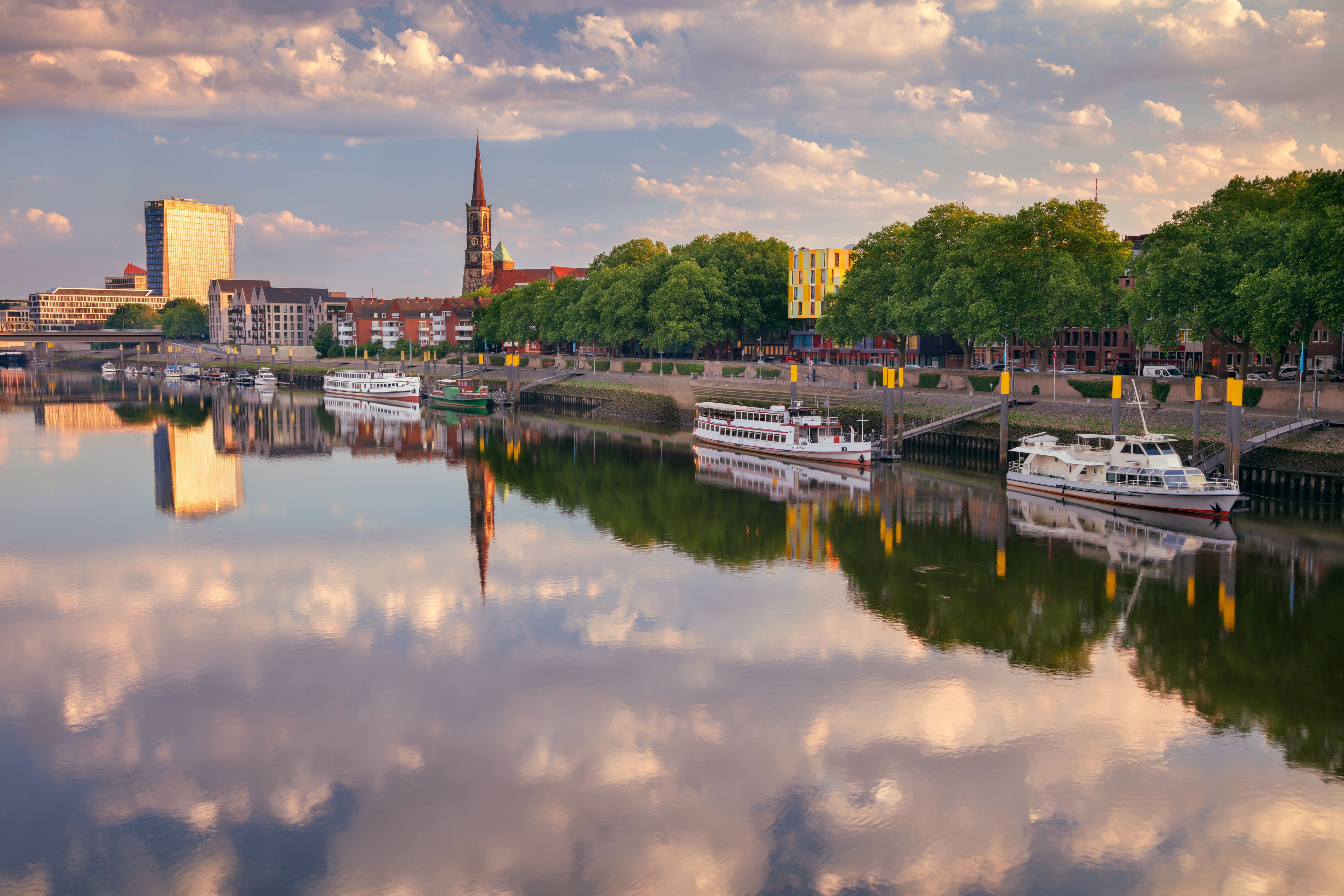 A calm river with boats, a church tower, and modern buildings along the riverbank.