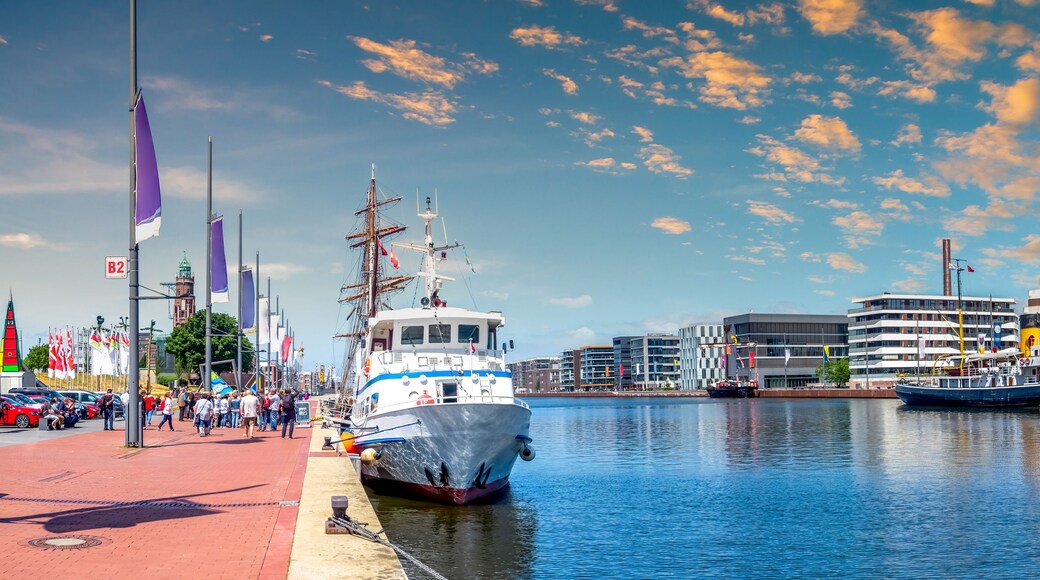 A harbor with a large sailing ship docked, a modern building complex, and a clear blue sky with scattered clouds.