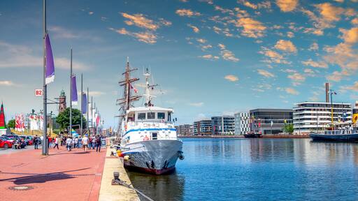 A harbor with a large sailing ship docked, a modern building complex, and a clear blue sky with scattered clouds.