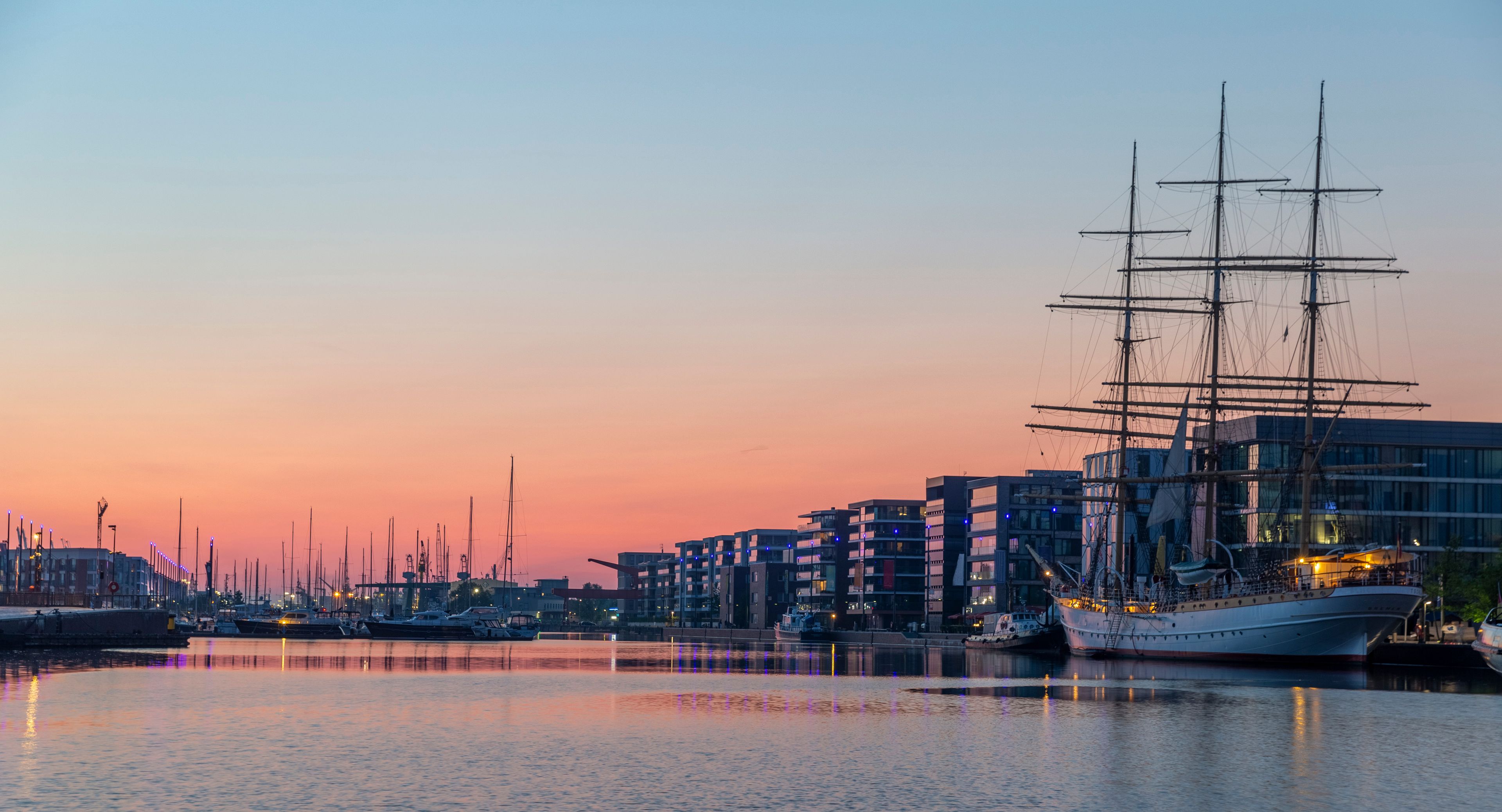 A harbor at sunset with a tall sailing ship docked, modern buildings, and multiple boats.
