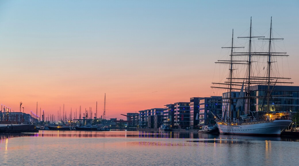 A harbor at sunset with a tall sailing ship docked, modern buildings, and multiple boats.