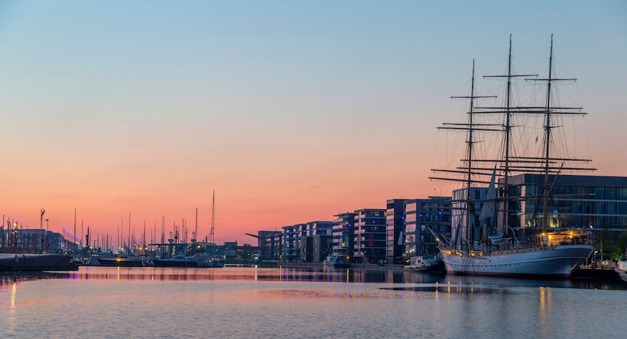 A harbor at sunset with a tall sailing ship docked, modern buildings, and multiple boats.