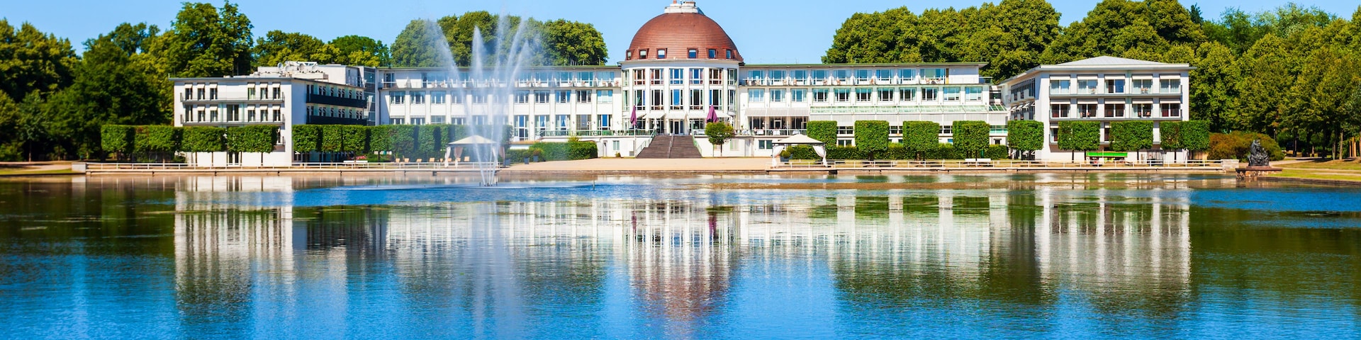 Un grand bâtiment surmonté d’un dôme, avec une fontaine et un lac paisible dans lequel se reflète la structure.