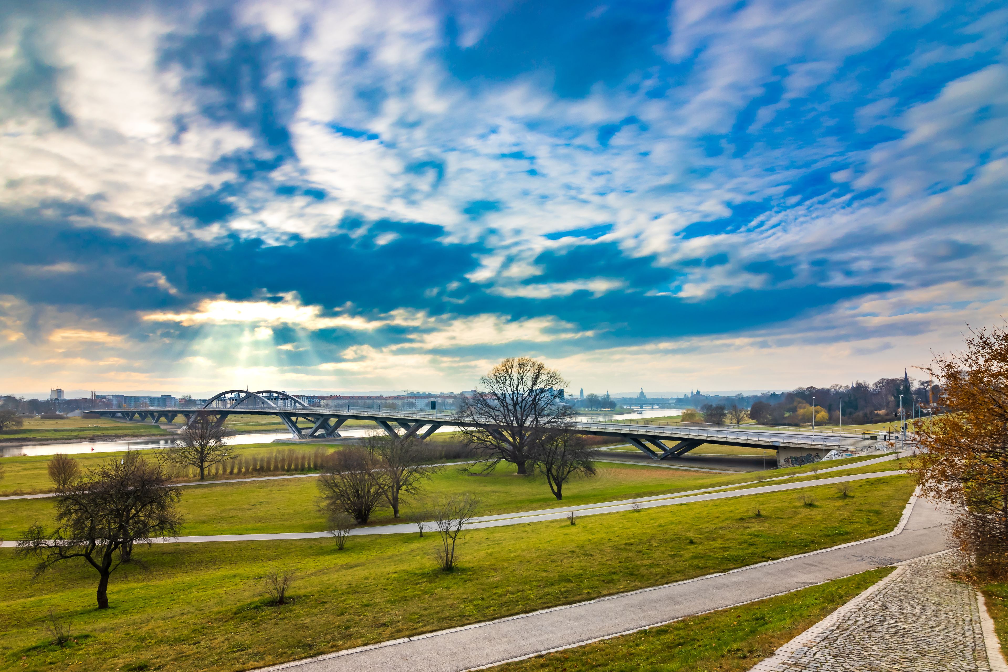 Eine Brücke über einem Fluss, im Vordergrund ein Park und im Hintergrund eine Stadtansicht.