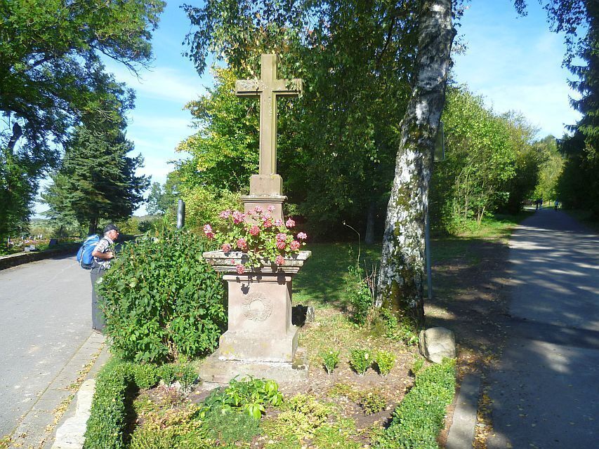 A stone cross with flowers, surrounded by greenery and trees.
