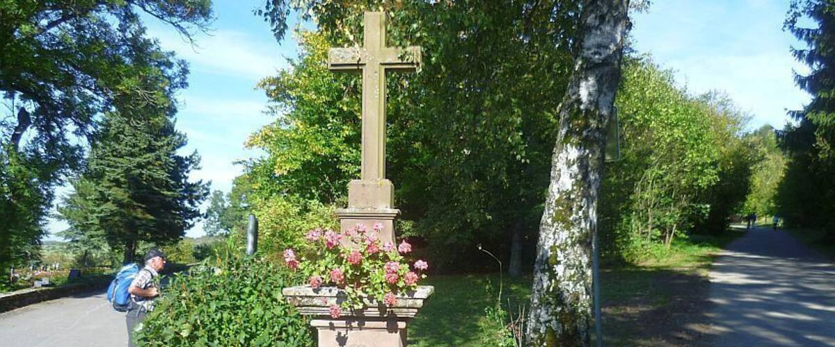 A stone cross with flowers, surrounded by greenery and trees.