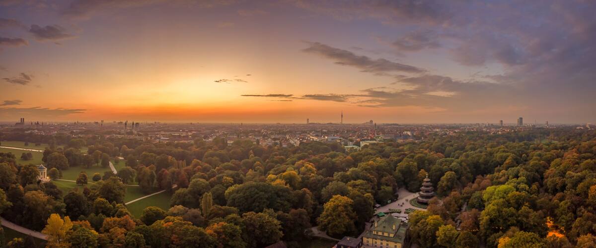 Ein weiter Panoramablick auf eine Stadt bei Sonnenuntergang, mit dichtem Wald im Vordergrund und einer Skyline im Hintergrund.