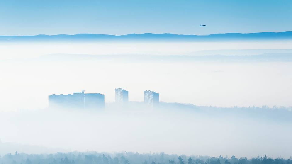 Un paisaje urbano con dos edificios destacados, cielo azul despejado y un avión volando.