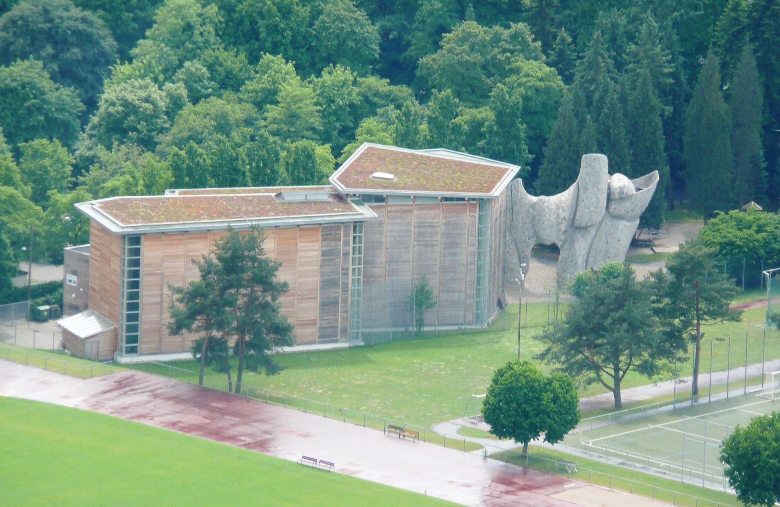 Un edificio moderno con una estructura singular que asemeja una roca, rodeado de frondosos árboles verdes y un campo deportivo.