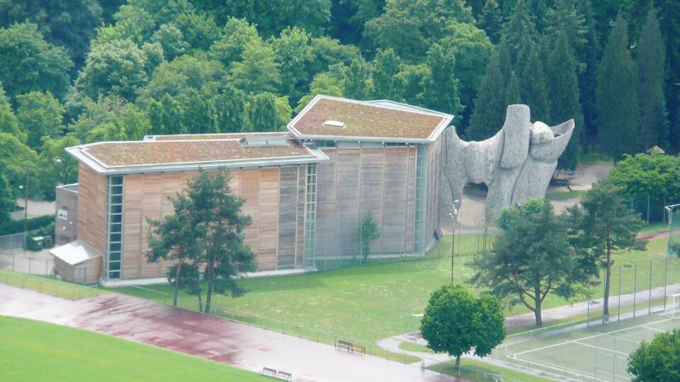 Un edificio moderno con una estructura singular que asemeja una roca, rodeado de frondosos árboles verdes y un campo deportivo.