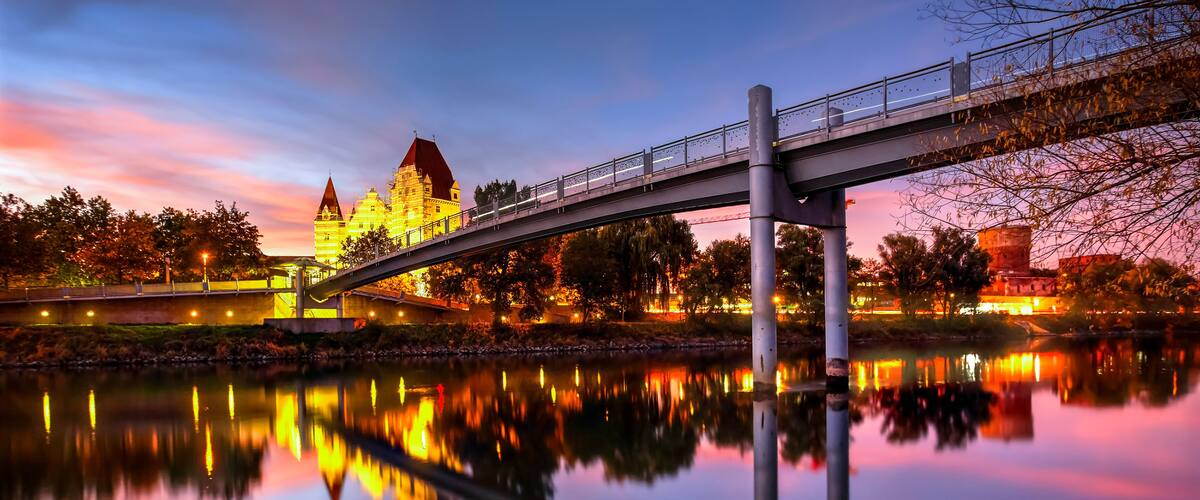 Eine Brücke über einem ruhigen Fluss, der das Licht eines Schlosses und der umliegenden Bäume widerspiegelt.