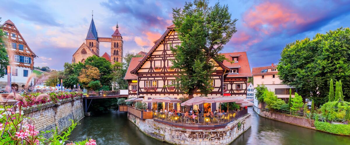 Eine Stadt am Flussufer mit traditionellen Fachwerkhäusern, einem Kirchturm und einer Brücke.