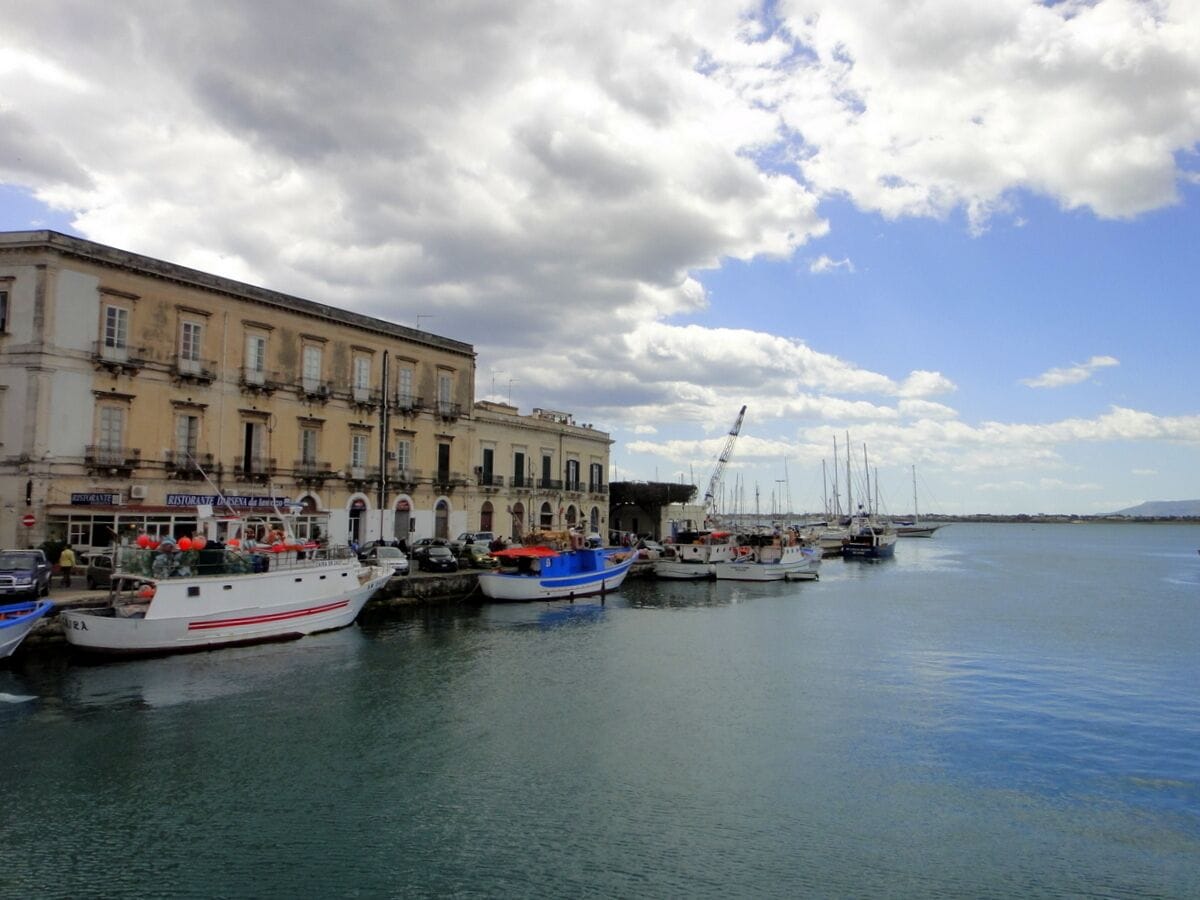 A harbor with several boats docked along the waterfront, a building with multiple stories and balconies, and a clear sky with scattered clouds.