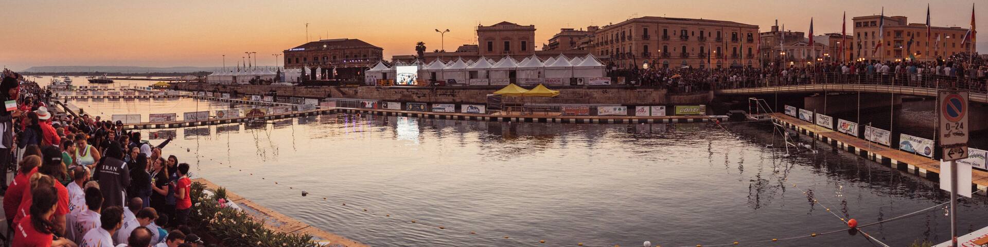 A waterfront with a crowd, buildings, and a sunset.