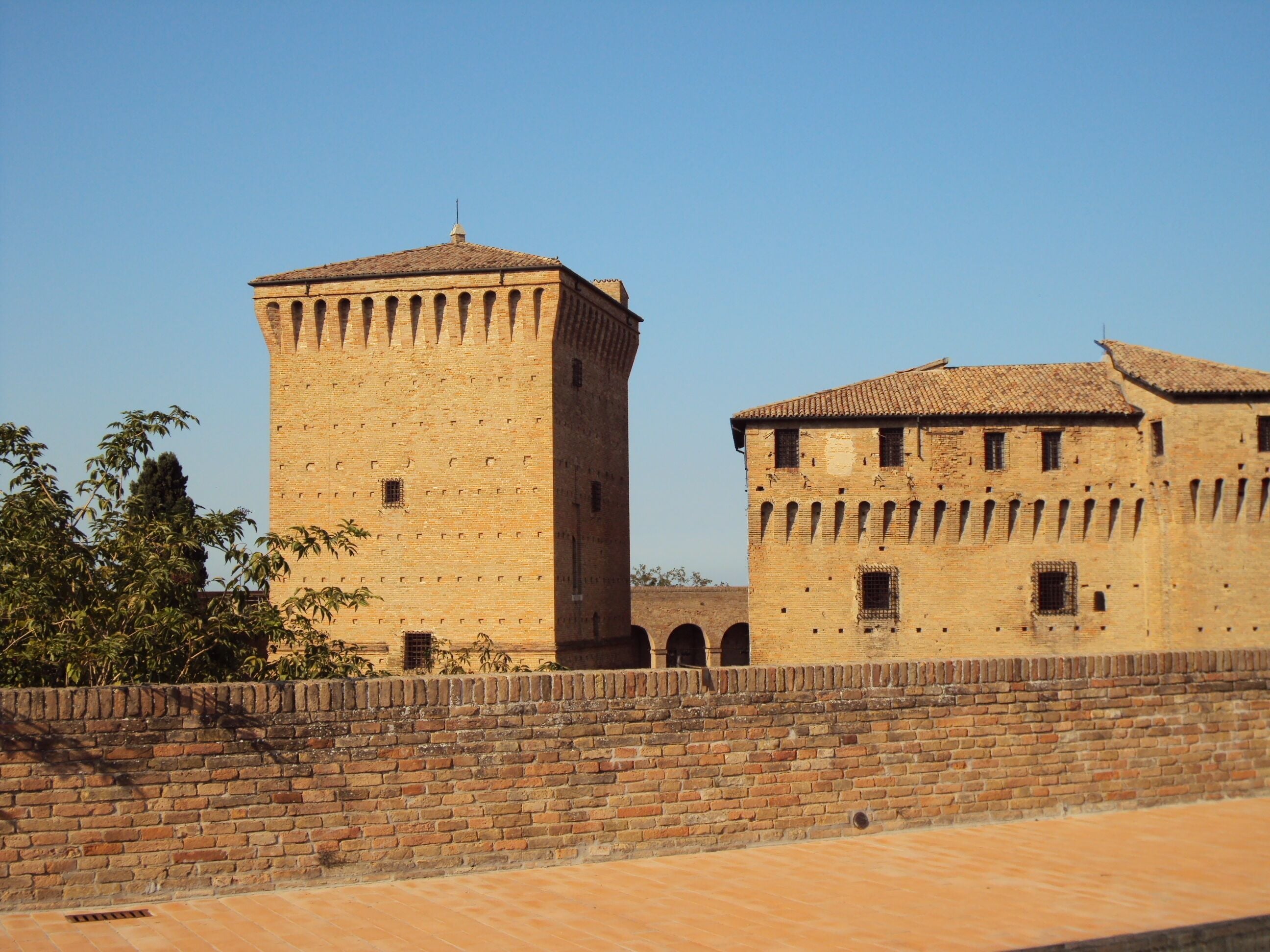Una torre de ladrillo histórica y un edificio con techo de tejas, rodeados por un muro de ladrillo y un sendero adoquinado.