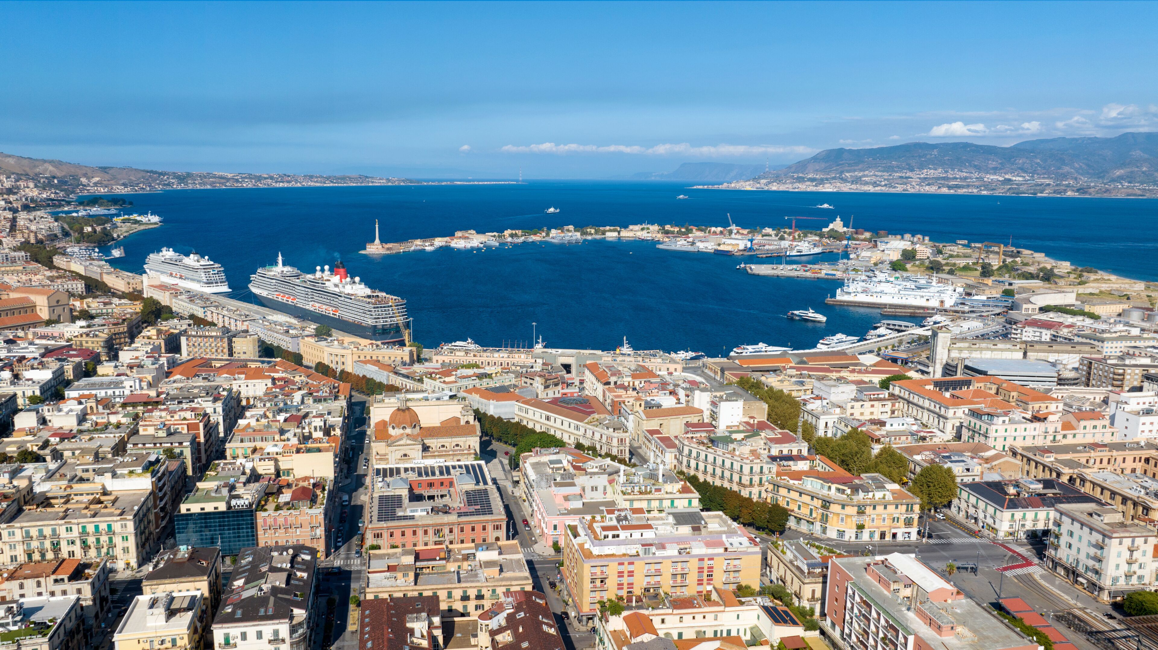 Aerial view of the city port of Messina, Sicily, Italy. Panorama of a crescent-shaped harbour. Large cruise ships and various vessels are docked along the quay. The historic center is in foreground.