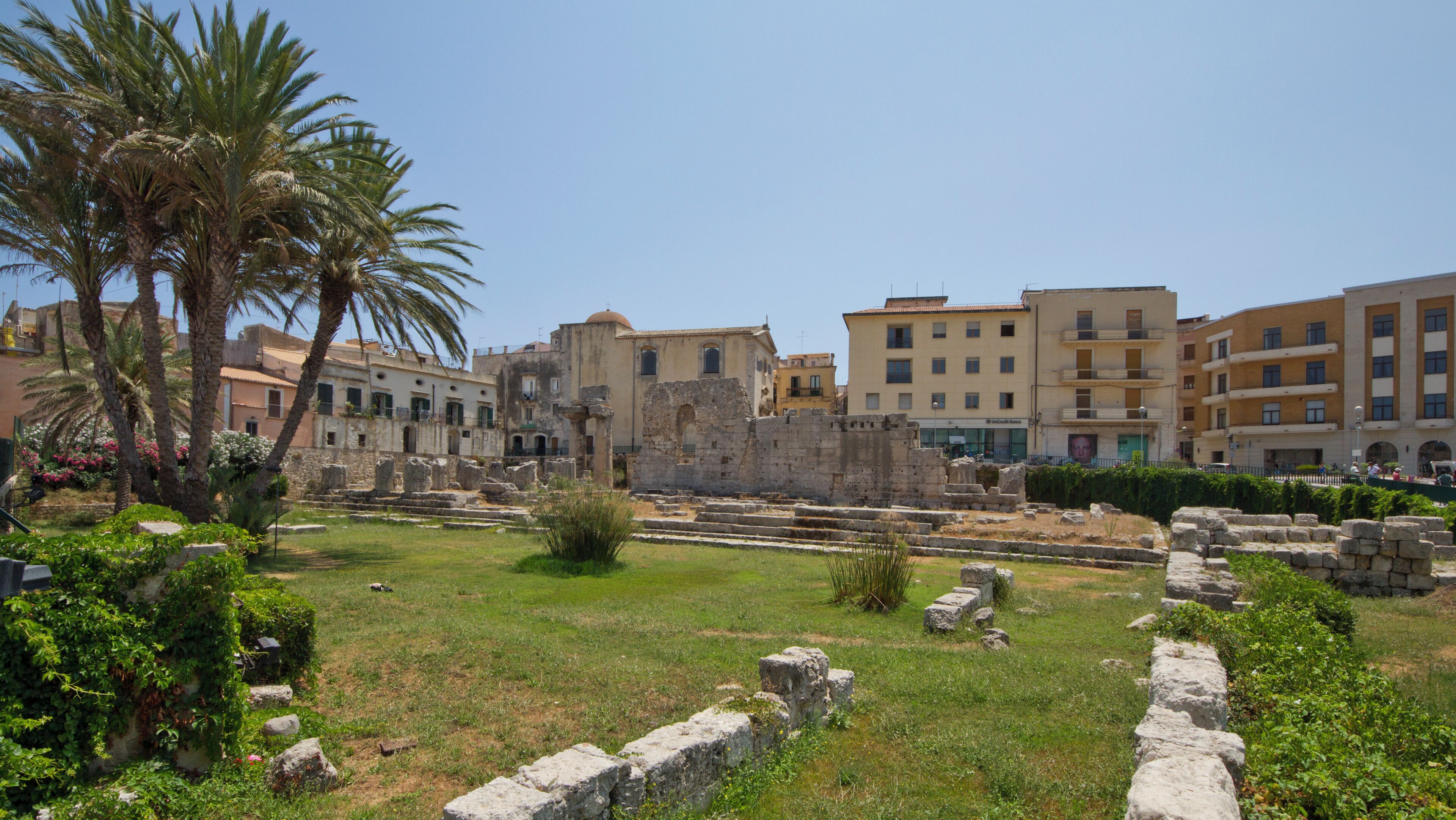 Ancient ruins with palm trees and modern buildings in the background.