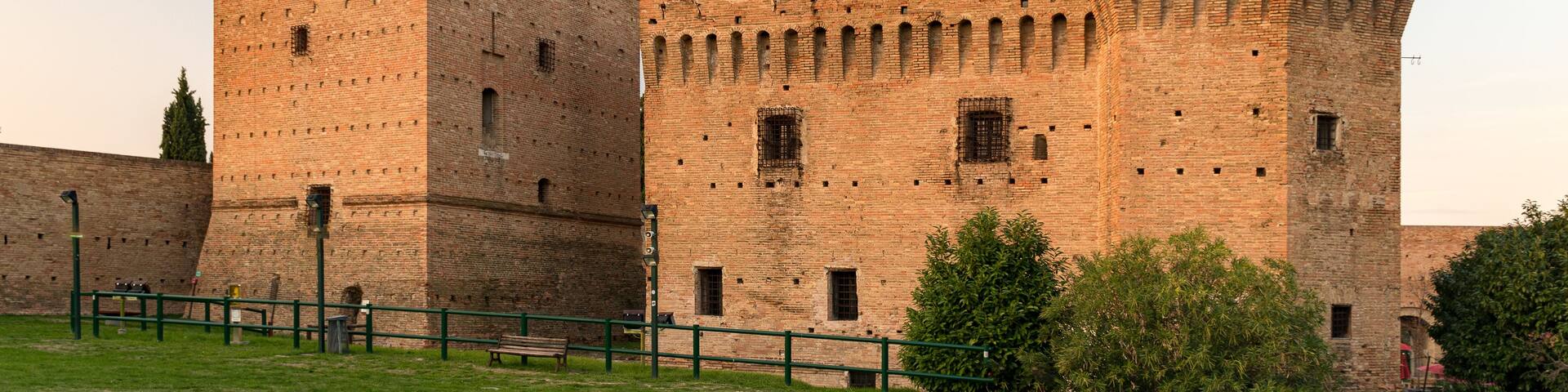 A historic brick fortress with two prominent towers, a grassy foreground, and a clear sky.