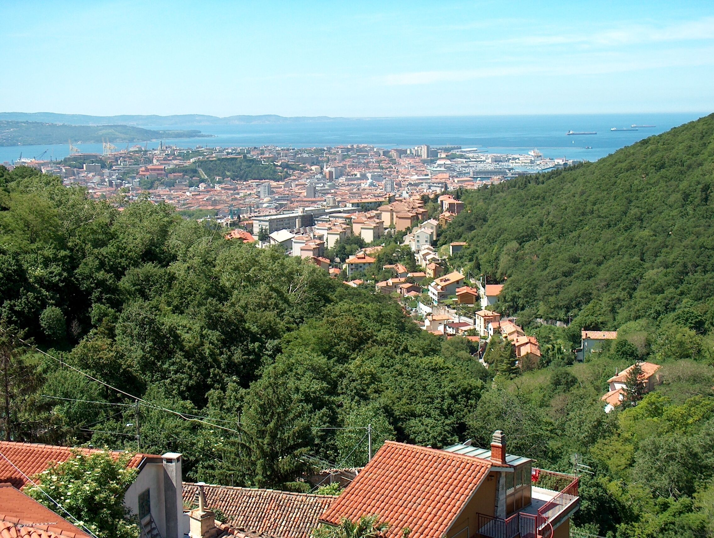 A coastal city with terracotta-roofed buildings nestled in a green valley, overlooking a calm sea and distant mountains.
