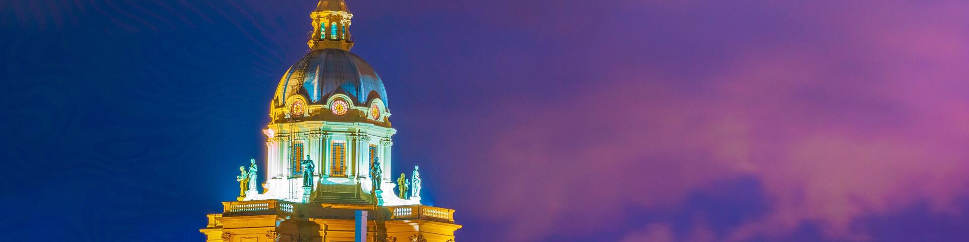 A grand, illuminated building with a large dome and statues, surrounded by trees and other buildings.