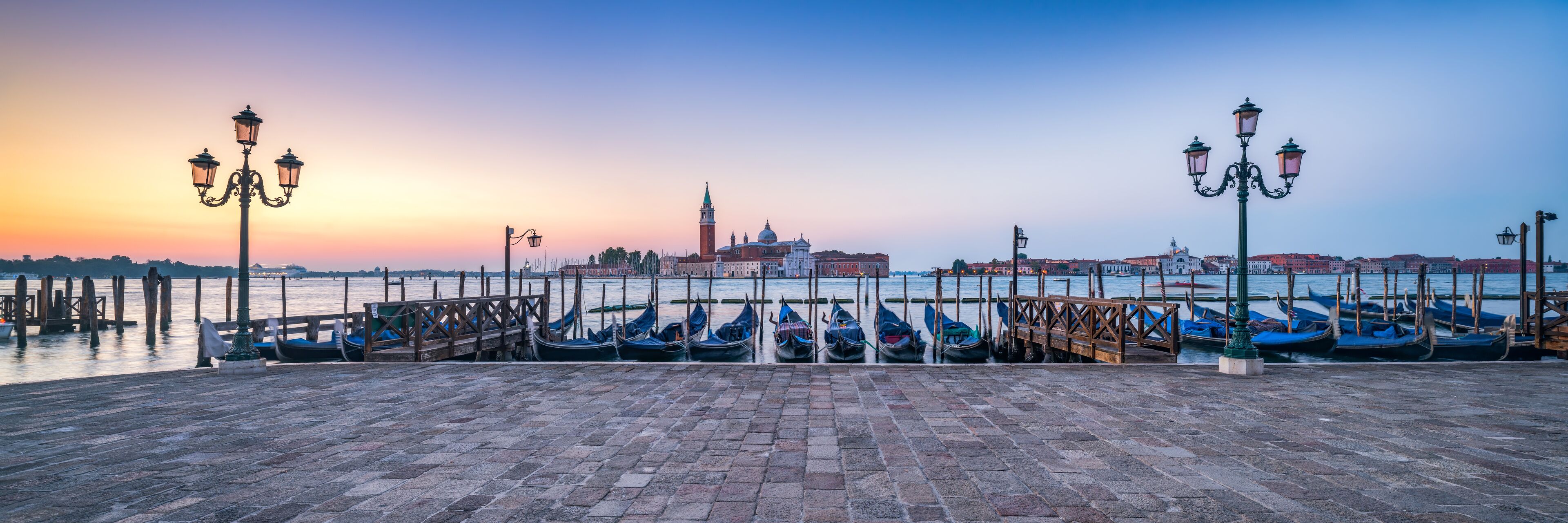 Une promenade au bord de l’eau où sont amarrées des gondoles, un bâtiment historique doté d’un dôme et d’une tour, et un ciel au coucher du soleil.