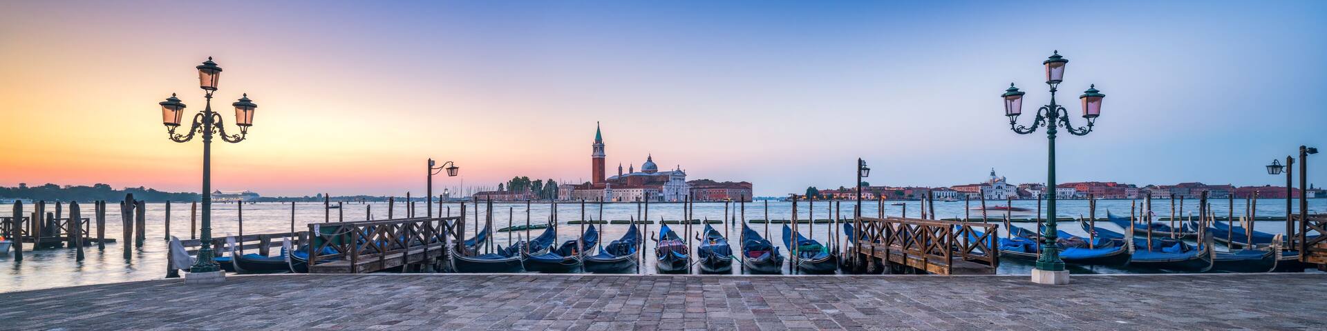 A waterfront with gondolas docked at a pier, a historic building with a dome and tower, and a sunset sky.