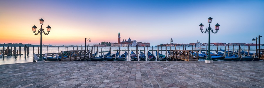 Une promenade au bord de l’eau où sont amarrées des gondoles, un bâtiment historique doté d’un dôme et d’une tour, et un ciel au coucher du soleil.