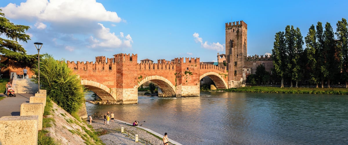 A historic stone bridge with arches over a river, a tall tower, and a paved walkway with people.