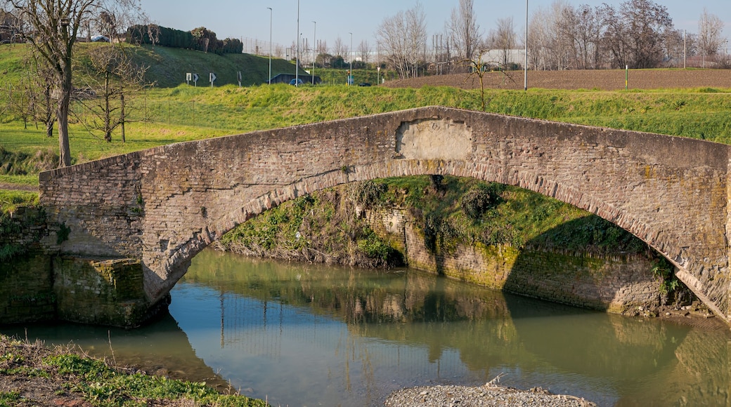 A stone bridge over a calm river, surrounded by greenery and fields.