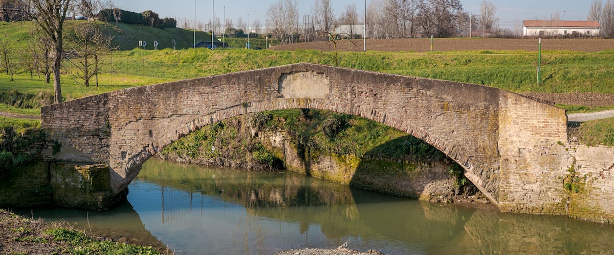 A stone bridge over a calm river, surrounded by greenery and fields.