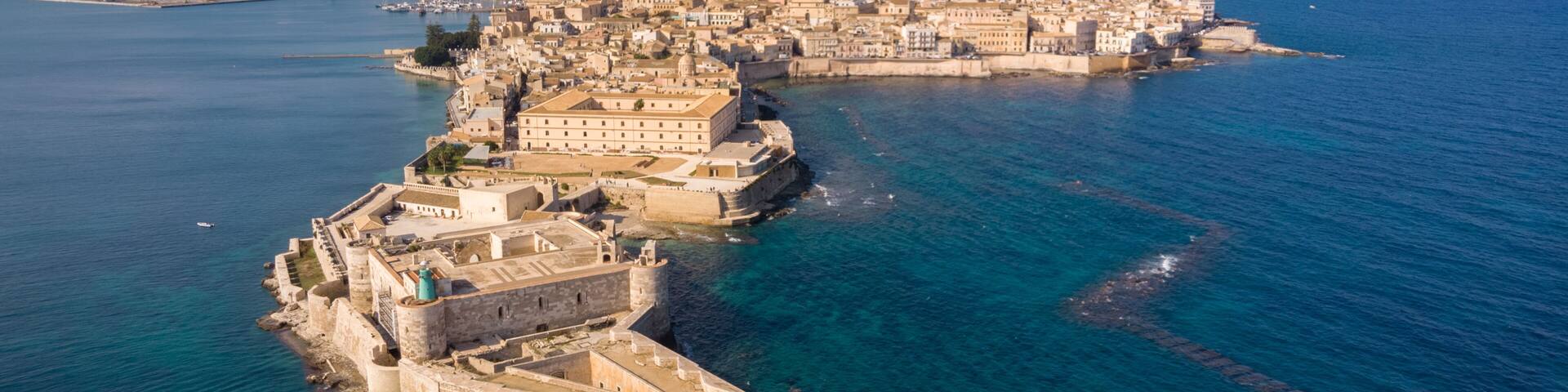 An aerial view of a coastal city with a prominent fortified structure and a densely built urban area in the background.