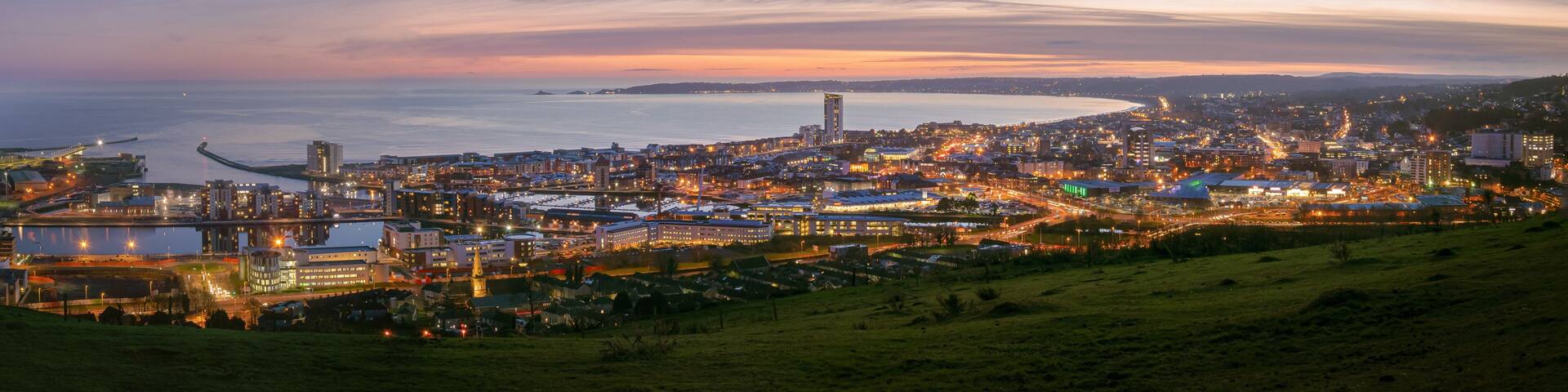 A coastal cityscape at dusk with illuminated buildings, a prominent church steeple, and a distant peninsula.