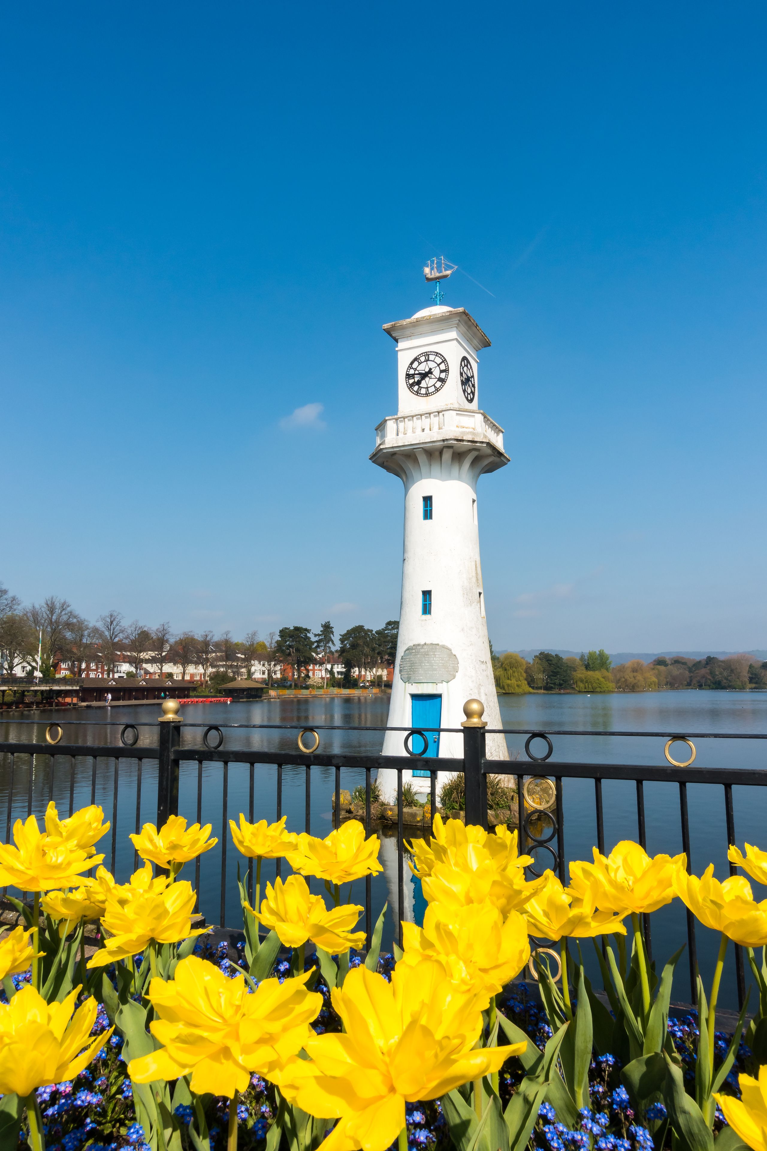 A white lighthouse with a clock tower, a blue door, and a plaque, surrounded by yellow flowers and a calm body of water.