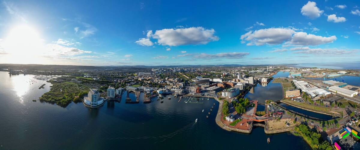 Aerial 180° Panorama Drone Shot of Cardiff Bay, Cardiff Bay Barrage, and Coastal Industrial Port with City Skyline, Waterfront Buildings, and Sunlight Reflections Under Blue Sky and Scattered Clouds,