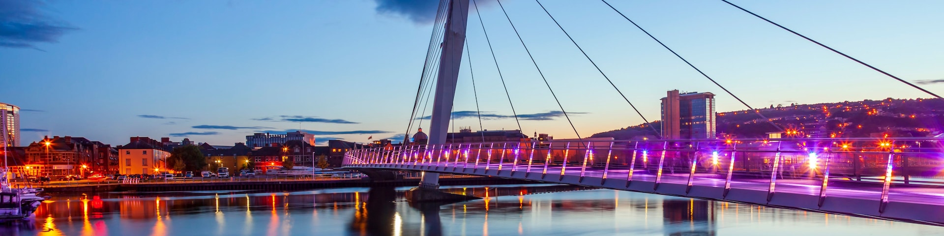 Uma ponte com cabos suspensos ao entardecer, com grades iluminadas refletindo na água.