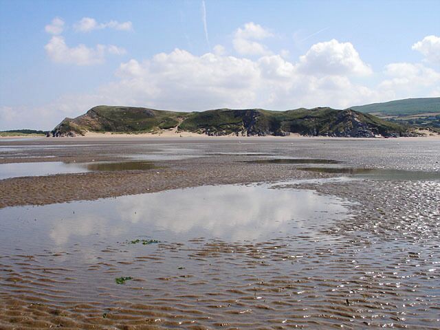 A coastal landscape with a sandy beach, shallow water, and grassy hills in the background.