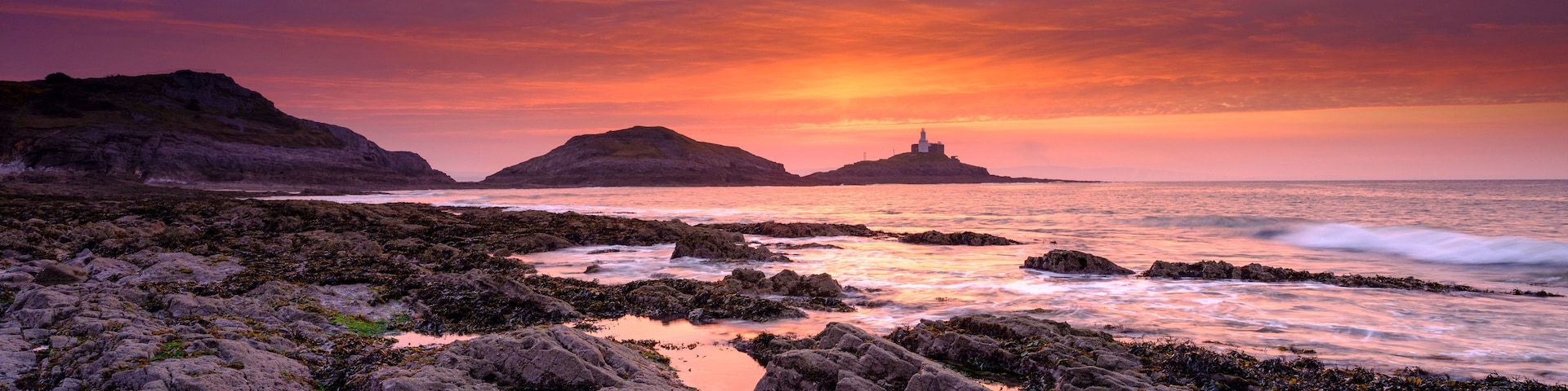 Uma costa rochosa com ondas quebrando nas pedras, um pôr do sol com cores vibrantes e um farol numa pequena ilha ao longe.