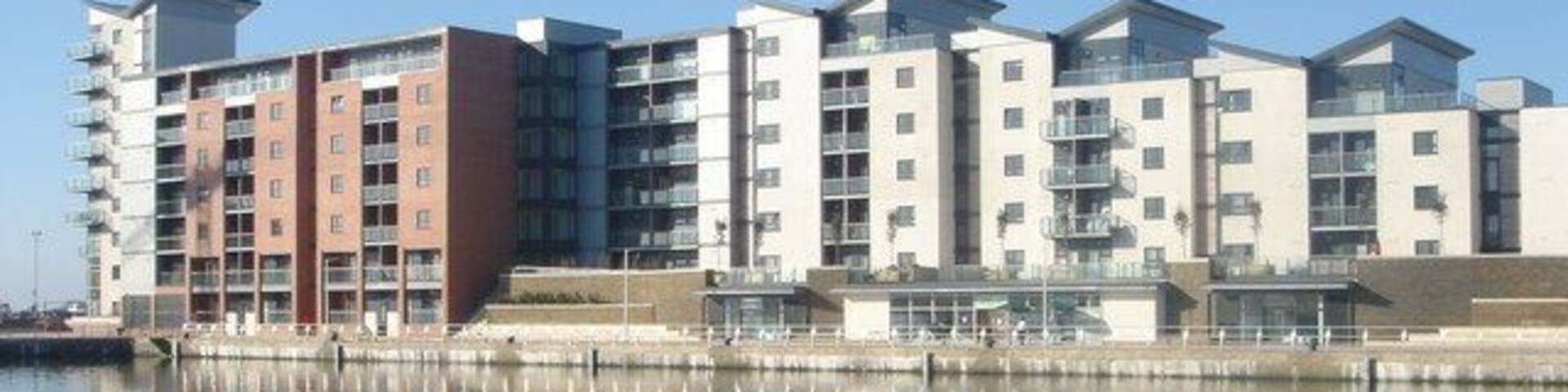 A waterfront residential building complex with multiple balconies and a calm body of water in the foreground.