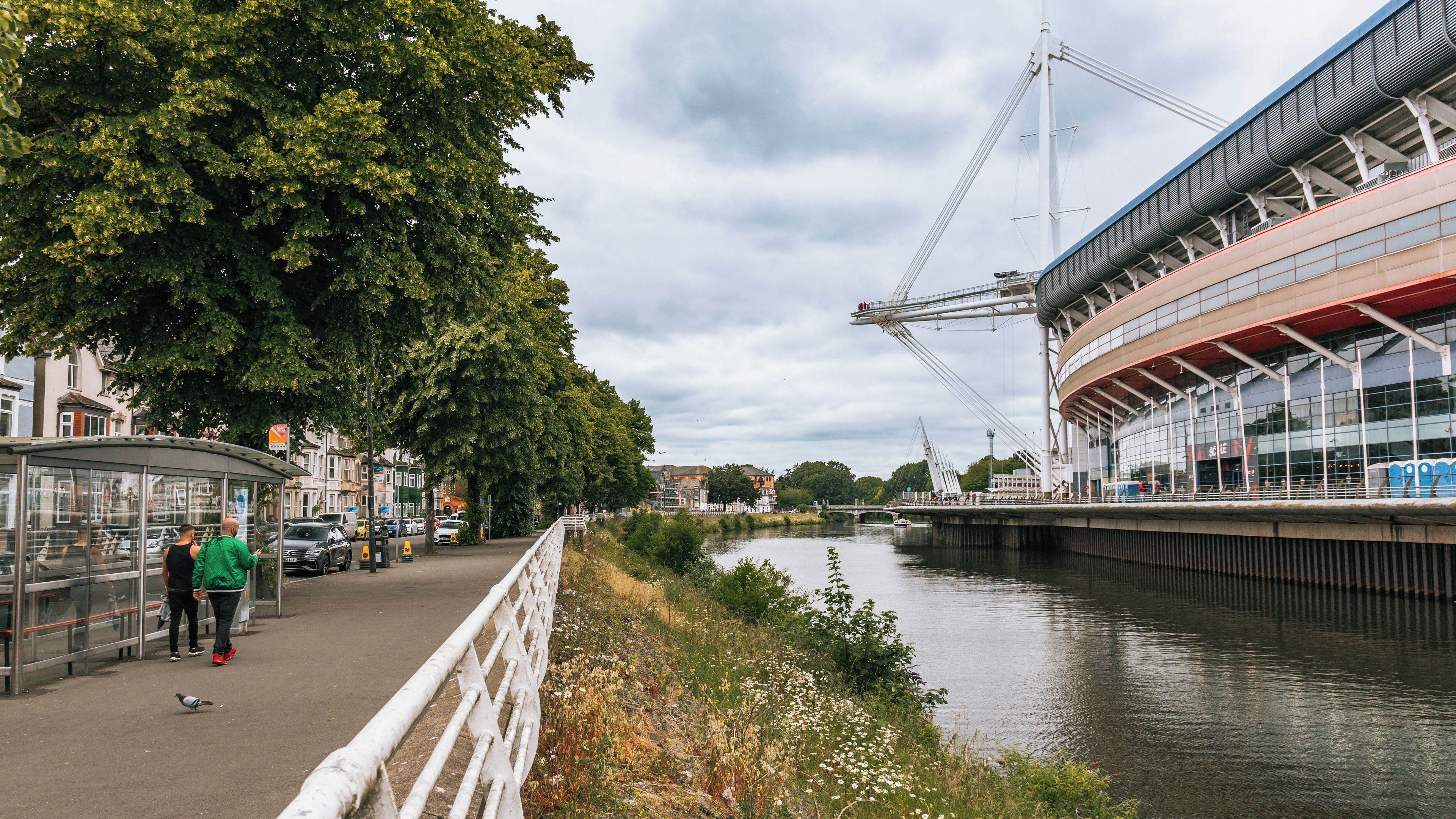 Un paseo fluvial con un puente peatonal, un estadio y árboles.
