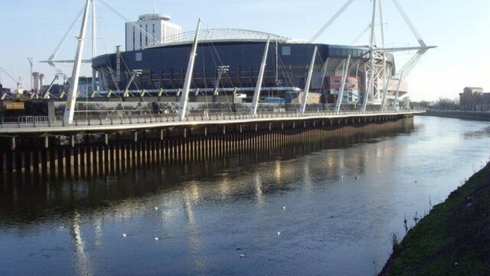 Ein modernes Stadion mit einer markanten Dachkonstruktion, ein Fluss im Vordergrund und eine grüne Böschung.