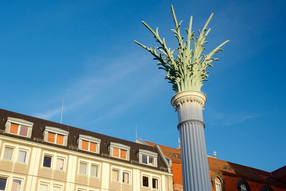 A tall, ornate column with a leafy top against a clear blue sky, with a multi-story building in the foreground.